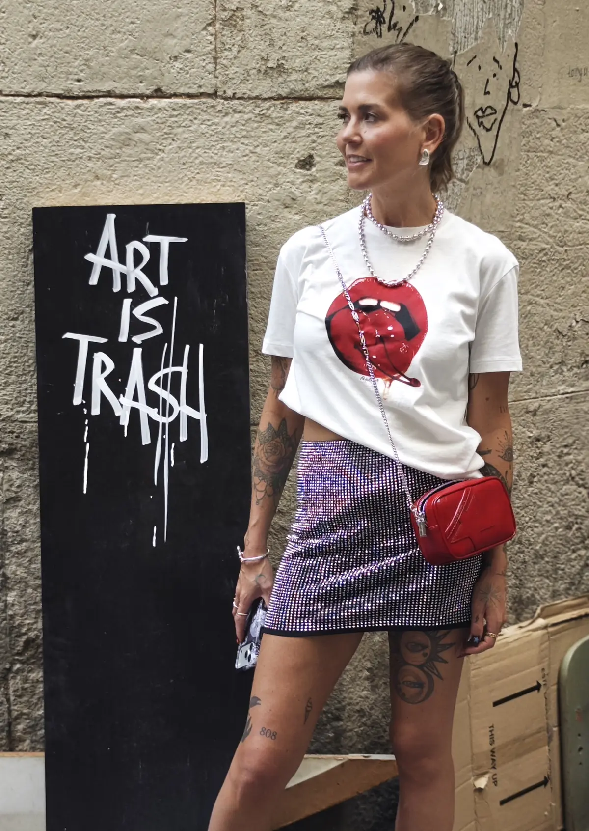 Cortnie leaning on a black door painted with the words Art Is Trash, wearing a red lips tee
